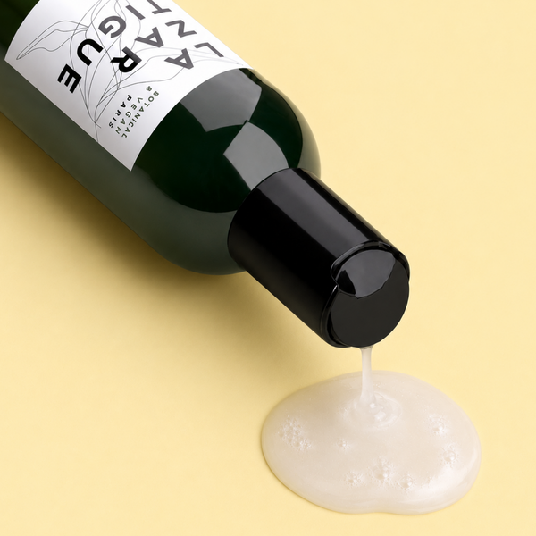 Lazartigue – Repair Shampoo (Intensiv reparierendes Shampoo), Nahaufnahme einer dunkelgrünen Shampooflasche, das Produkt fließt auf eine gelbe Oberfläche