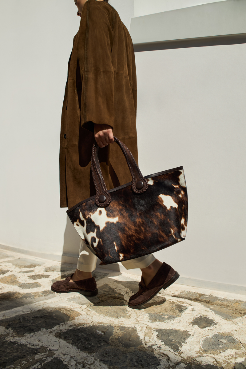 A model walking outdoors holding a dark brown and cream cow print pattern tote bag with dark brown rope handles , wearing a long brown coat and white pants with dark brown shoes.