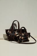 A dark brown and cream cow print pattern medium tote bag with dark brown rope handles and a small tote bag in the same color, texture and shape with dark brown rope handles and strap on a light gray background.