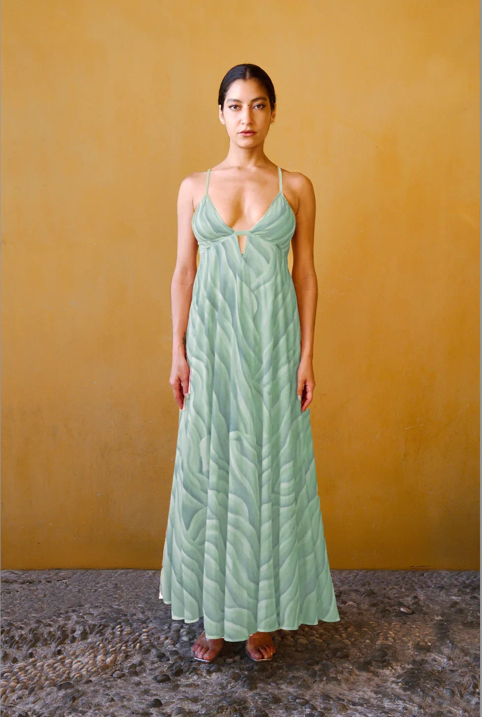 Mujer con vestido largo estampado verde claro de tirantes finos, de pie frente a una pared color mostaza