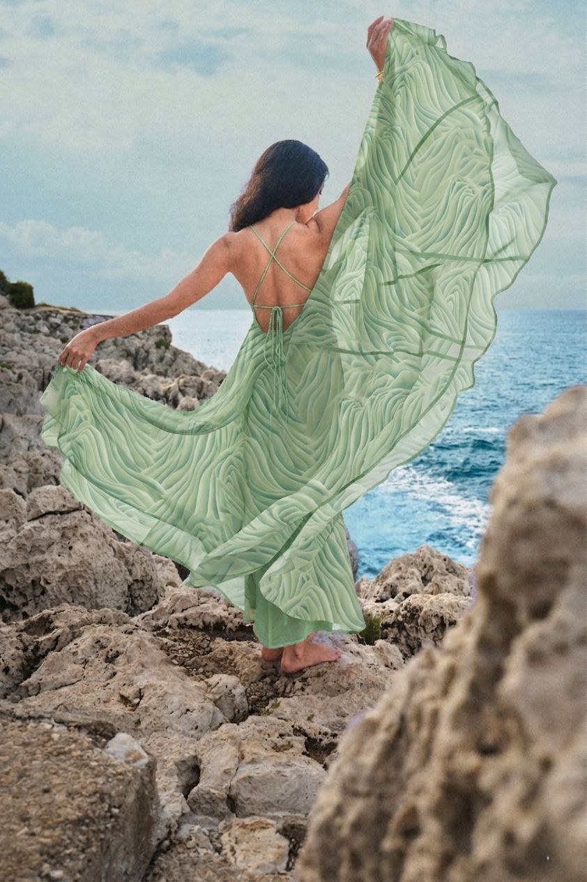 Mujer con vestido verde fluido de pie en una costa rocosa con vistas al océano