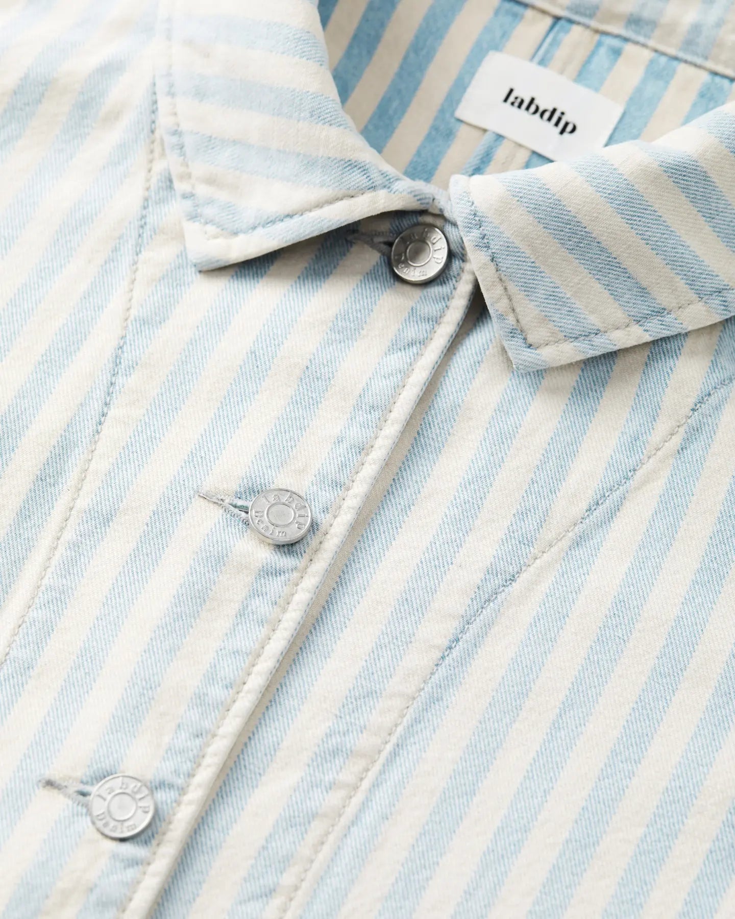 Détail d'une veste en denim léger rayé bleu ciel et écru, avec boutons métalliques gravés Labdip, tissu en coton structuré et coupe ajustée.