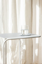 Beistelltisch DENYA mit Glas Wasser darauf in einem hellen Raum vor Vorhängen.