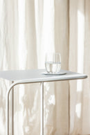Beistelltisch DENYA mit Glas Wasser darauf in einem hellen Raum vor Vorhängen.