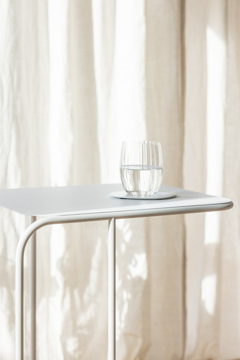 Beistelltisch DENYA mit Glas Wasser darauf in einem hellen Raum vor Vorhängen.