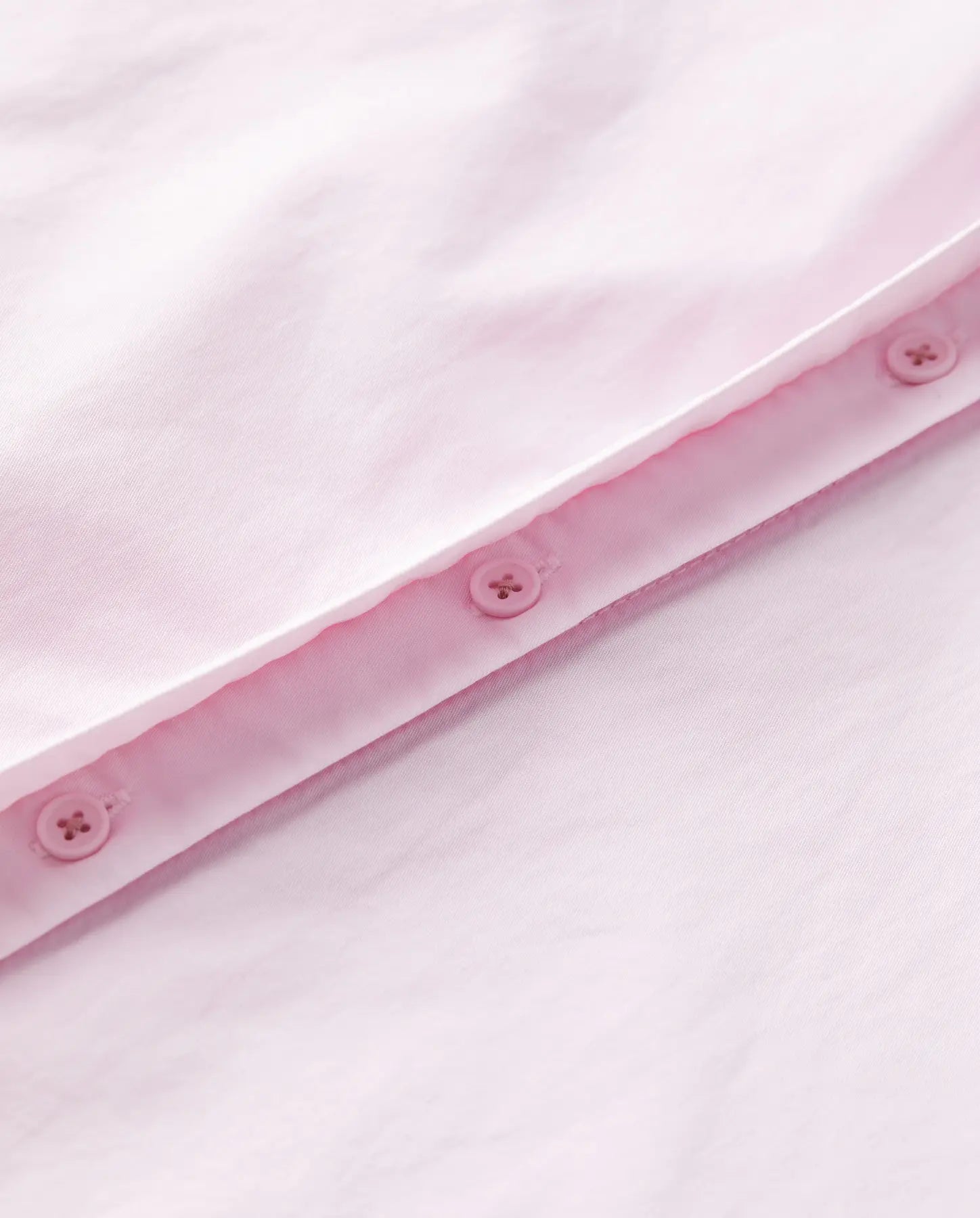 Détail d’un tissu popeline rose clair en coton, lisse et léger, avec une finition soignée comprenant des boutons assortis cousus sur une couture propre.