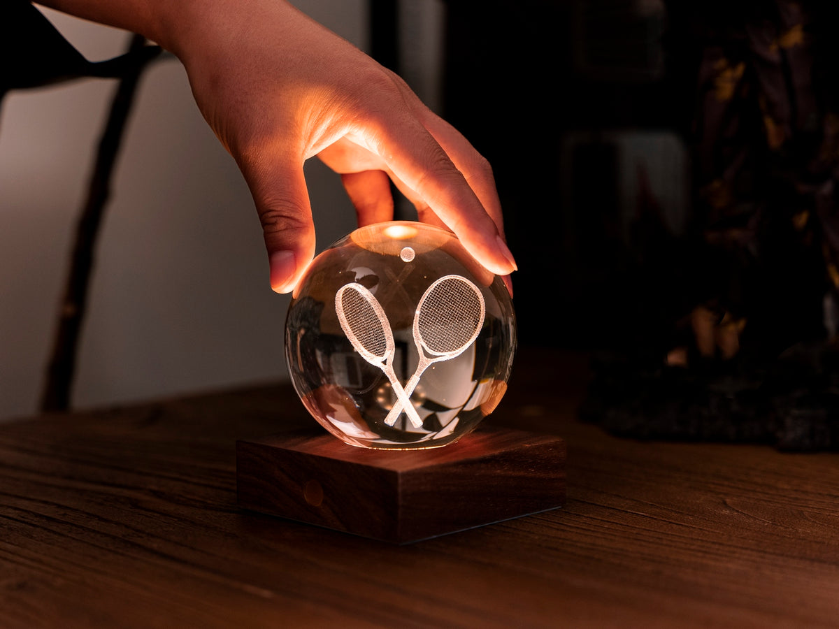 Gingko Design – Amber Crystal Light - Tennisracket, een hand raakt een oplichtende kristallen bol op een houten voet aan