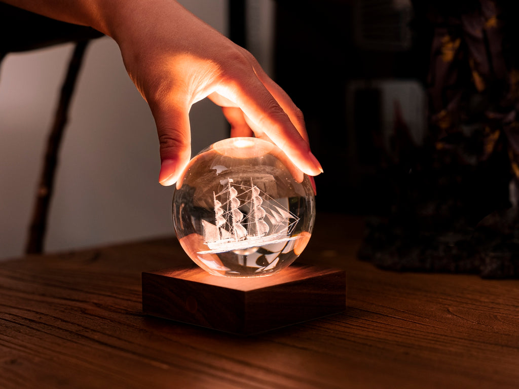 Gingko Design – Lampka Amber Crystal - Żaglówka, dłoń dotykająca świecącej szklanej kuli z grawerowanym statkiem