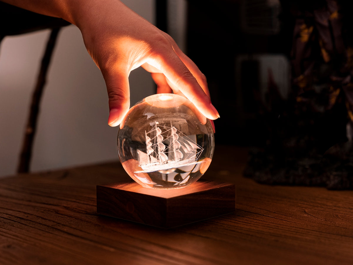 Gingko Design – Lampka Amber Crystal - Żaglówka, dłoń dotykająca świecącej szklanej kuli z grawerowanym statkiem