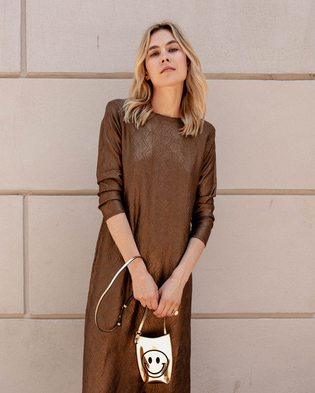 Michonet – Abito Foil Cobre, modella bionda con abito marrone, con borsa dorata smiley