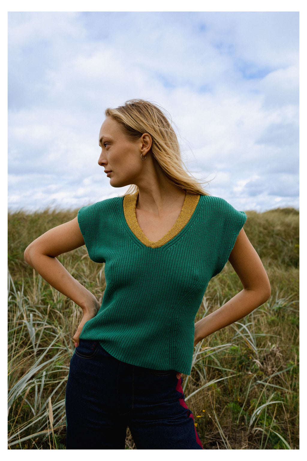 Michonet – Ärmlös Väst Knitted Green, modell i grön tröja och blå byxor, poserande i profil i gräset