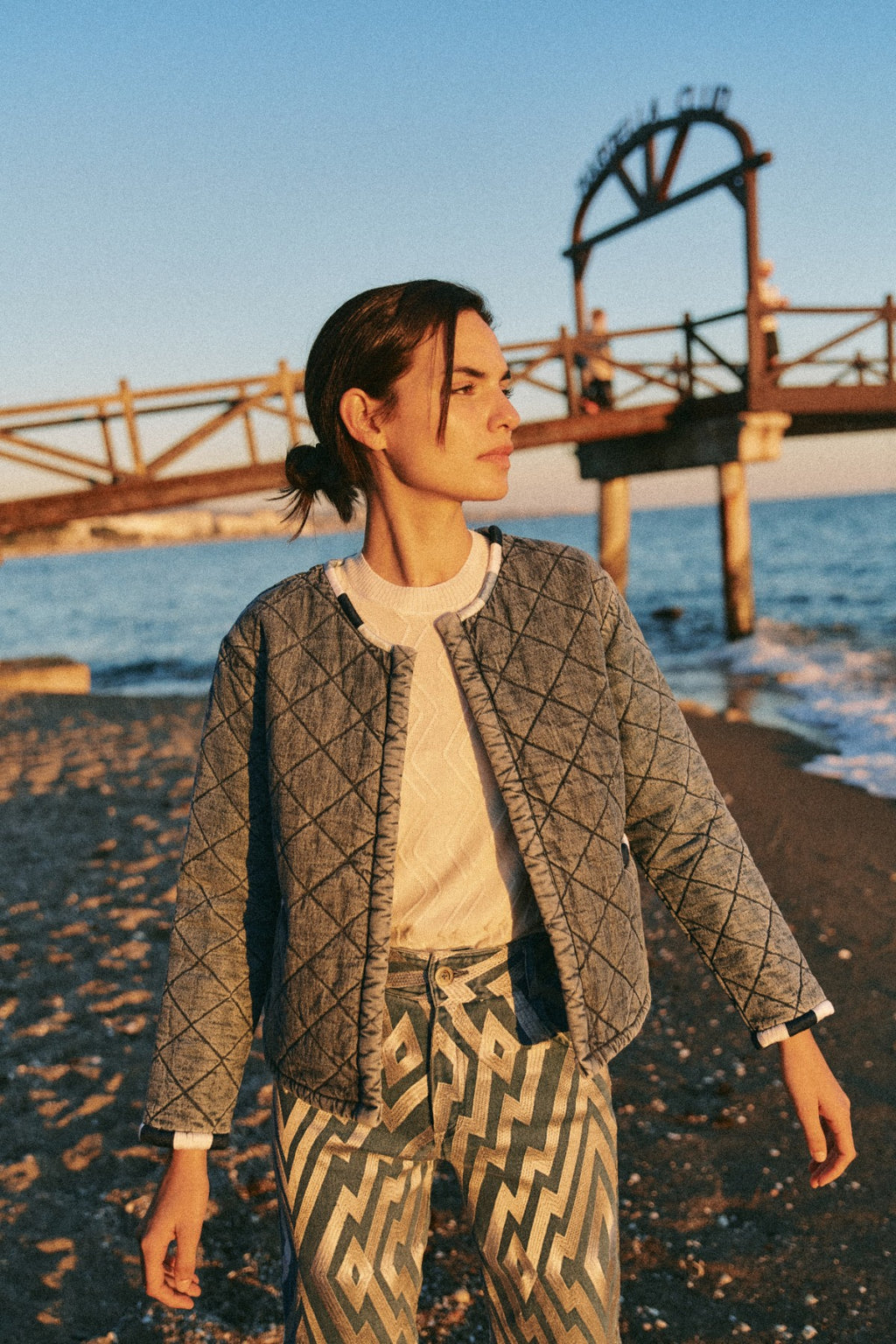 Michonet – Veste Denim Guata, mannequin de profil portant une veste matelassée grise sur la plage