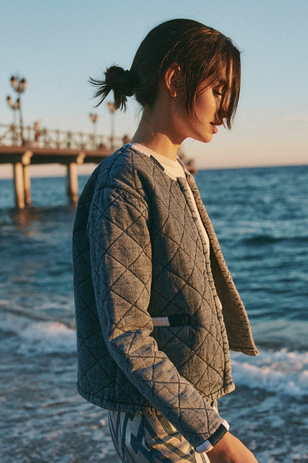 Michonet – Veste Denim Guata, mannequin de profil portant une veste matelassée grise sur la plage