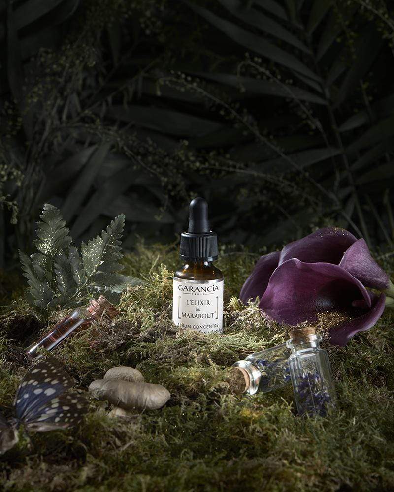 Garancia Marabout Elixir Serum presented in an enchanted forest setting with moss, purple flowers, a butterfly and magic vials.