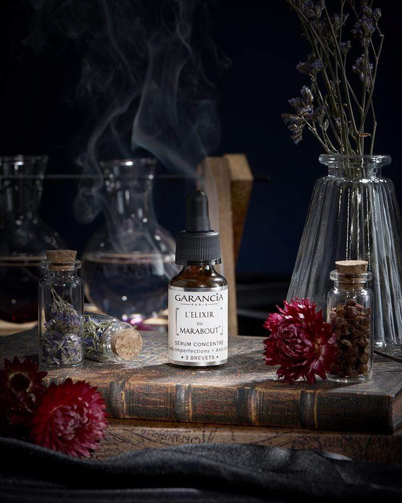 L'Élixir du Marabout de Garancia serum placed on an old grimoire, surrounded by red flowers, apothecary vials and a mystical smoke evoking a witchcraft laboratory.