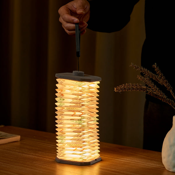 Gingko Design – Lámpara Smart Accordion - Terciopelo Gris Landmark, una persona sostiene una linterna de papel plisado y luminosa sobre una mesa de madera