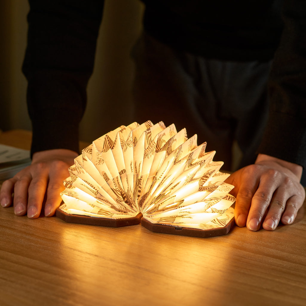 Gingko Design – Lampe Smart Accordion - Velours Marron Zèbre, lampe livre ouverte sur une table en bois, mains d'une personne