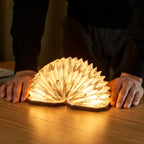 Gingko Design – Lampe Smart Accordion - Velours Marron Zèbre, lampe livre ouverte sur une table en bois, mains d'une personne