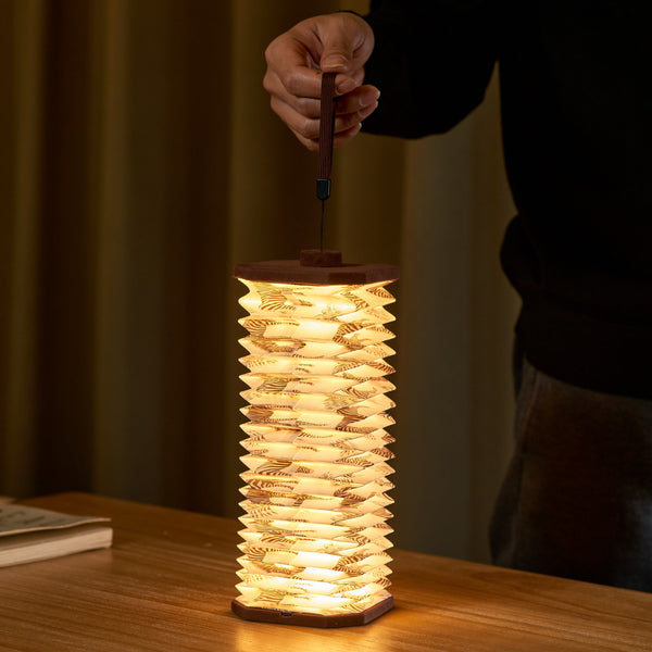 Gingko Design – Lámpara Smart Accordion - Terciopelo Marrón Cebra, una persona sostiene una linterna de papel plisado iluminada sobre una mesa de madera