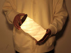 Gingko Design – Luz Twist Hexagon - Nogal Macizo, una persona sostiene una lámpara plegable de papel blanco luminoso