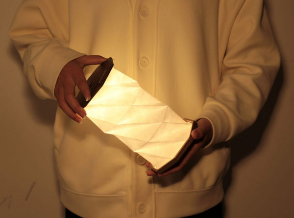 Gingko Design – Luz Twist Hexagon - Nogal Macizo, una persona sostiene una lámpara plegable de papel blanco luminoso