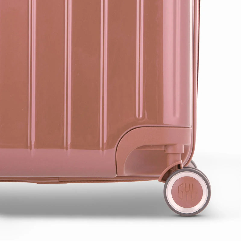 Detalle de la rueda y el logo GYL en la parte inferior de la maleta baúl de policarbonato brillante en color oro rosa. #color_oro rosa