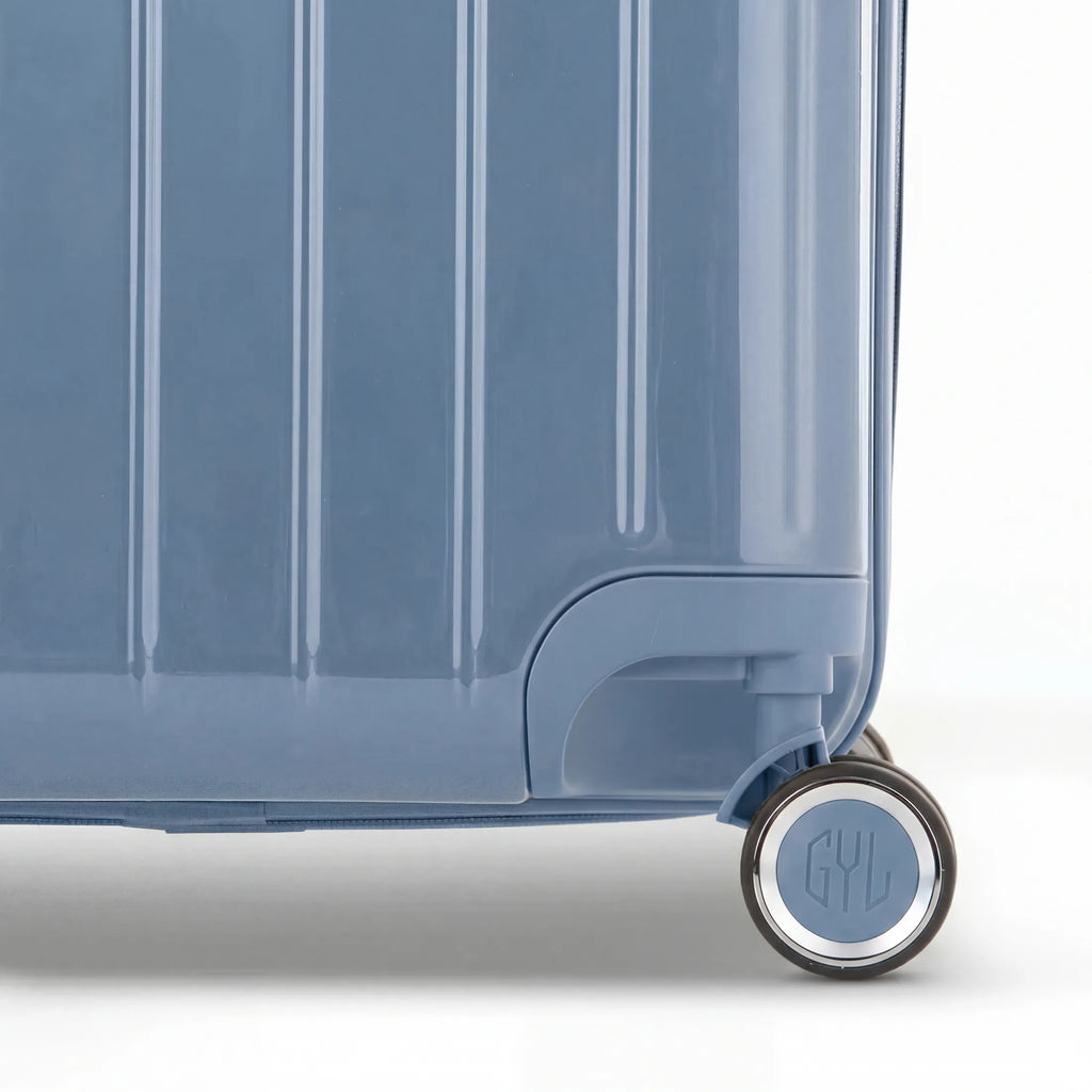 Détail de la roue et du logo GYL sur le bas de la valise trunk bleu gris en polycarbonate brillant. #couleur_bleu grisé