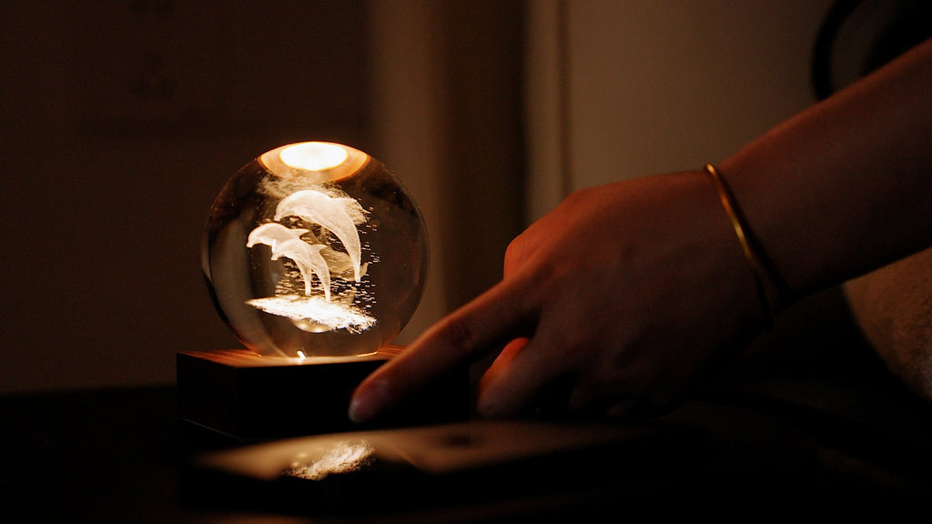 Gingko Design – Amber Crystal Leuchte - Delfine, eine Hand berührt eine leuchtende Glaskugel mit eingravierten Delfinen