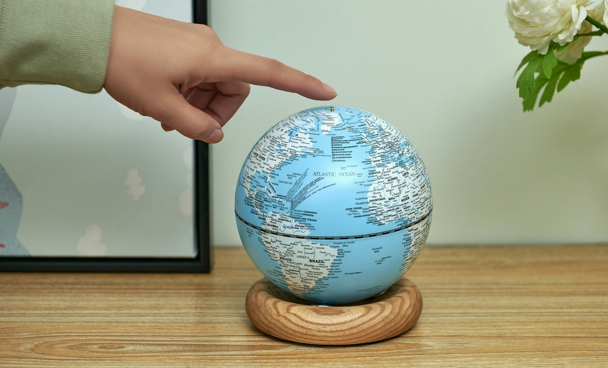 Gingko Design – Globuslampe Atlas - Hellblau / Sockel Weiße Esche - Mini, eine Hand berührt einen blauen Globus auf einem Holzsockel