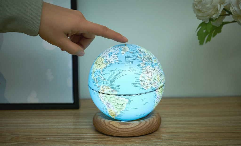 Gingko Design – Globuslampe Atlas - Hellblau / Sockel Weiße Esche - Mini, eine Hand berührt einen leuchtenden Globus auf einem Holzsockel