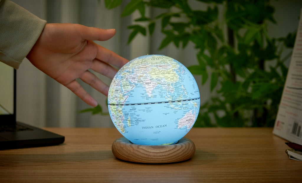 Gingko Design – Globuslampe Atlas - Hellblau / Sockel Weiße Esche - Mini, eine Hand berührt einen leuchtenden Globus auf einem Schreibtisch