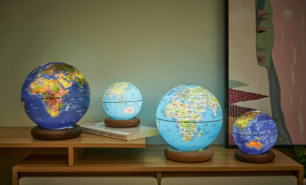 Gingko Design – Globuslampe Atlas - Hellblau / Sockel Weiße Esche - Mini, vier leuchtende Globen in verschiedenen Größen auf einem Holzmöbel