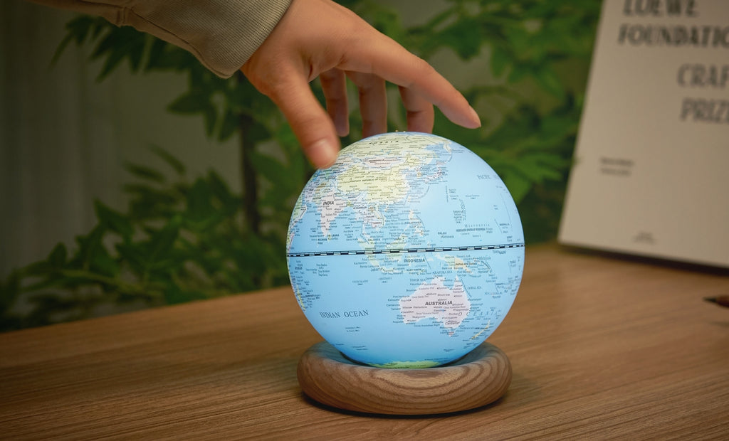 Gingko Design – Globuslampe Atlas - Hellblau / Sockel Weiße Esche - Mini, eine Hand berührt einen leuchtenden Globus auf einem Holzsockel