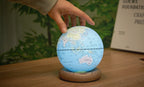 Gingko Design – Globuslampe Atlas - Hellblau / Sockel Weiße Esche - Mini, eine Hand berührt einen leuchtenden Globus auf einem Holzsockel