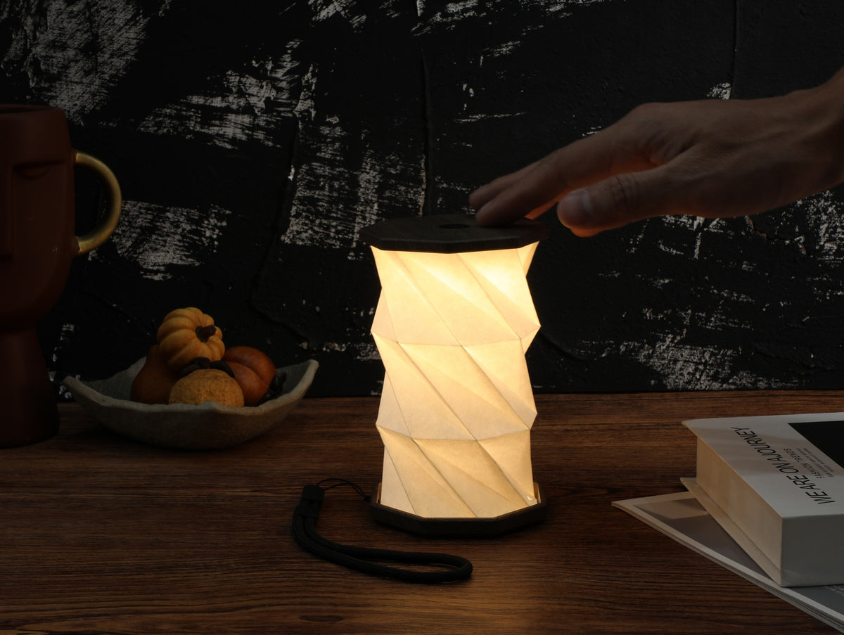 Gingko Design – Twist Hexagon Leuchte - Treibholz Walnuss - Mini, faltbare Tischleuchte eingeschaltet auf einem Holztisch platziert