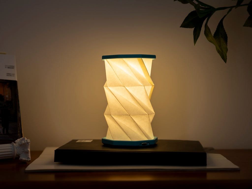 Gingko Design – Luz Twist Hexagon - Azul Laguna - Mini, lámpara de sobremesa plegable encendida, colocada sobre un libro, ambiente cálido