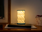 Gingko Design – Luz Twist Hexagon - Azul Laguna - Mini, lámpara de sobremesa plegable encendida sobre una mesa de madera