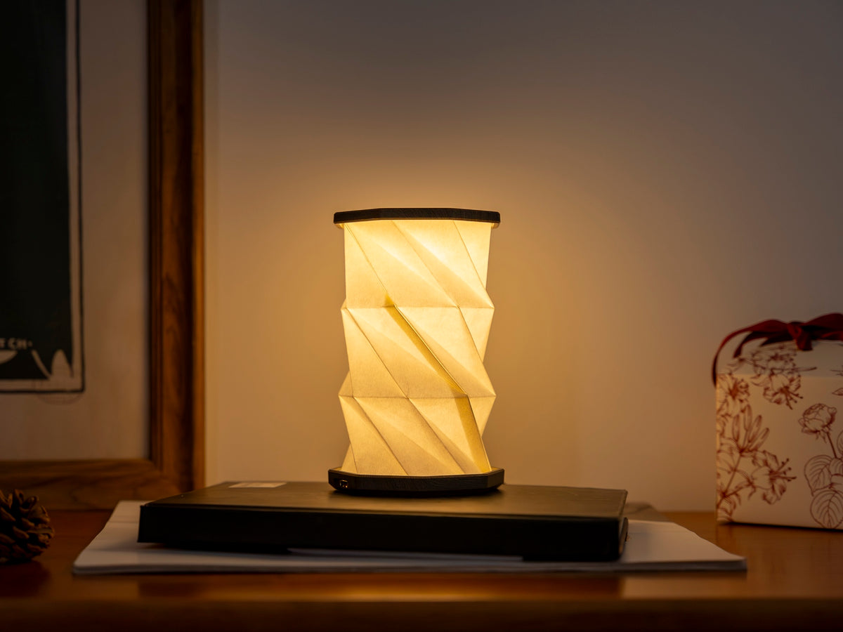 Gingko Design – Twist Hexagon Leuchte - Treibholz Walnuss - Mini, faltbare Tischleuchte aus weißem Papier eingeschaltet auf einem Holztisch