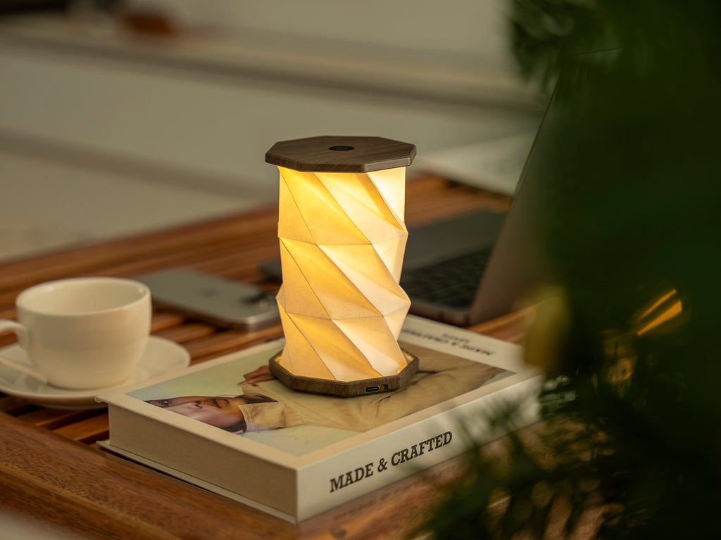 Gingko Design – Twist Hexagon Leuchte - Treibholz Walnuss - Mini, eingeschaltete Tischleuchte auf einem Buch, mit einer Tasse