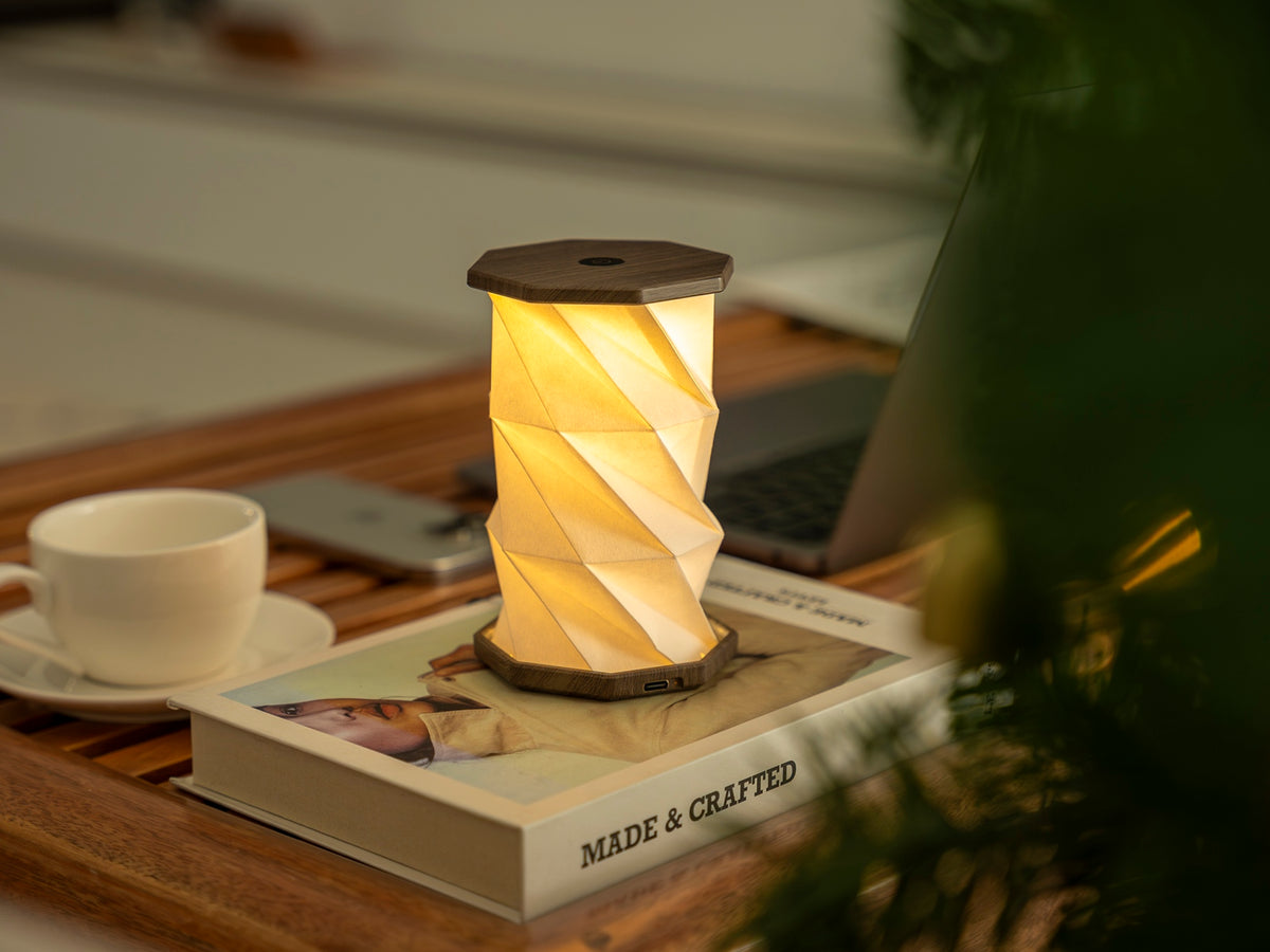 Gingko Design – Twist Hexagon Leuchte - Treibholz Walnuss - Mini, eingeschaltete Tischleuchte auf einem Buch, mit einer Tasse