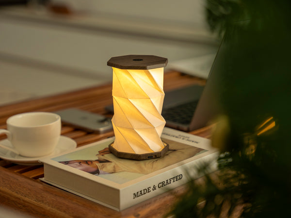 Gingko Design – Luz Twist Hexagon - Nogal Flotado - Mini, lámpara de sobremesa encendida sobre un libro, con una taza