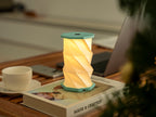 Gingko Design – Luz Twist Hexagon - Azul Laguna - Mini, lámpara de mesa encendida sobre un libro, con taza y ordenador