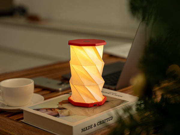 Gingko Design – Luz Twist Hexagon - Rojo Coral - Mini, lámpara de sobremesa encendida sobre un libro, con una taza