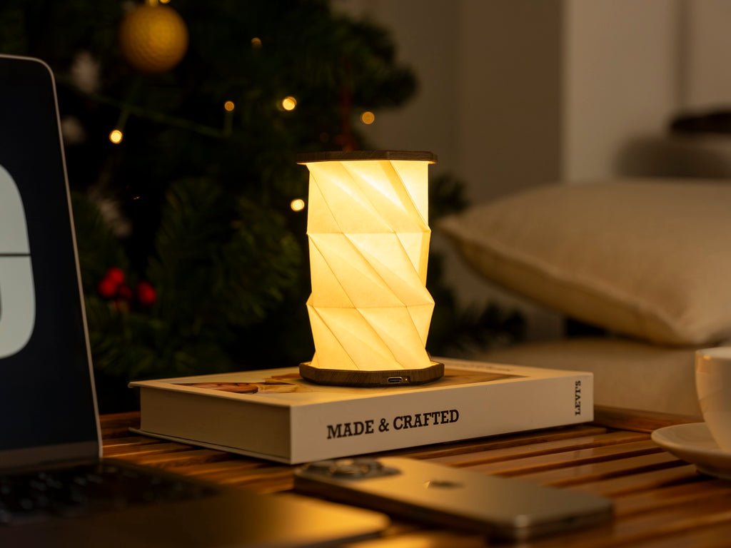 Gingko Design – Twist Hexagon Leuchte - Treibholz Walnuss - Mini, eingeschaltete Tischleuchte auf einem Buch, neben einem Laptop