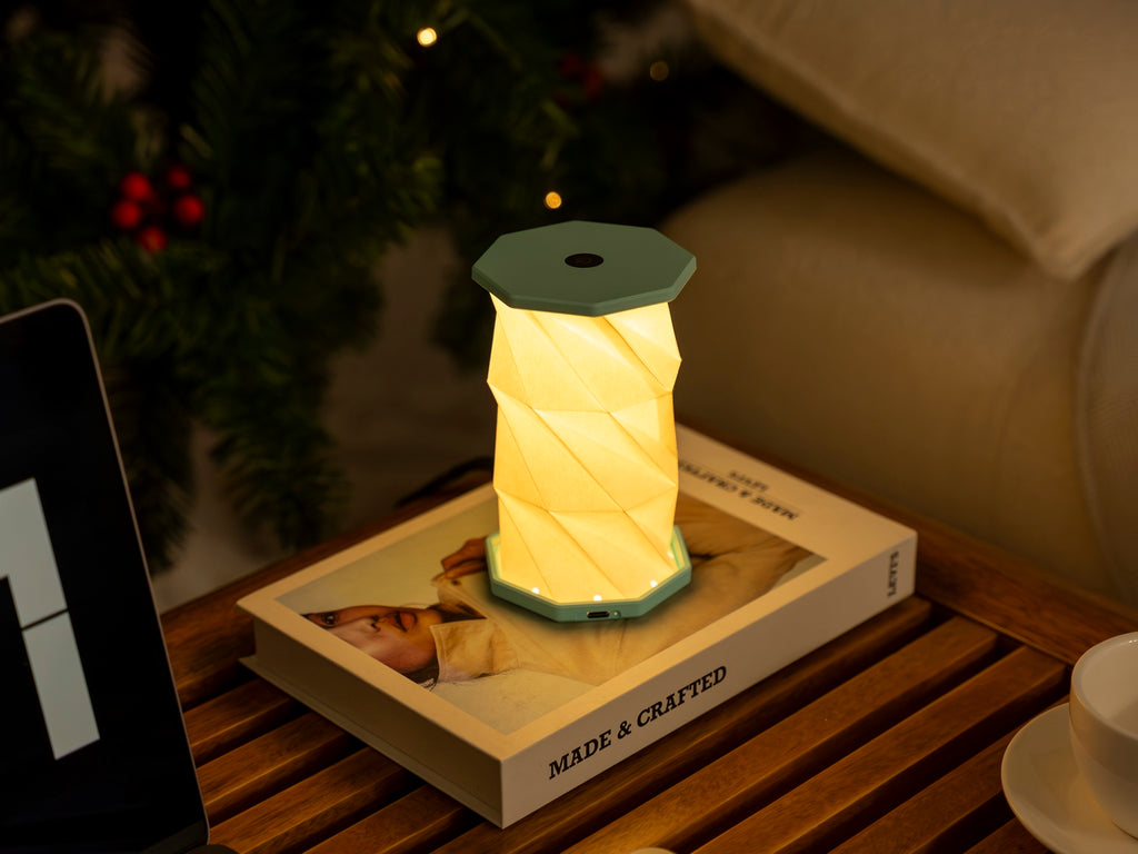 Gingko Design – Luz Twist Hexagon - Azul Laguna - Mini, lámpara de sobremesa plegable encendida sobre un libro, ambiente cálido