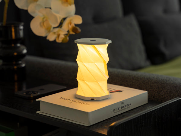 Gingko Design – Luz Twist Hexagon - Blanco Lunar - Mini, lámpara de mesa plegable encendida colocada sobre un libro blanco