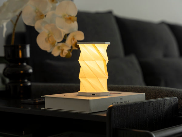 Gingko Design – Luz Twist Hexagon - Blanco Lunar - Mini, lámpara de mesa encendida sobre un libro, en un salón oscuro