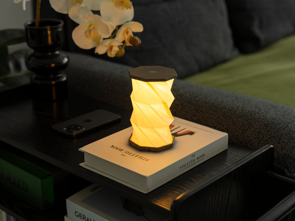 Gingko Design – Twist Hexagon Leuchte - Treibholz Walnuss - Mini, eingeschaltete Tischleuchte auf einem Buch in einem gemütlichen Interieur