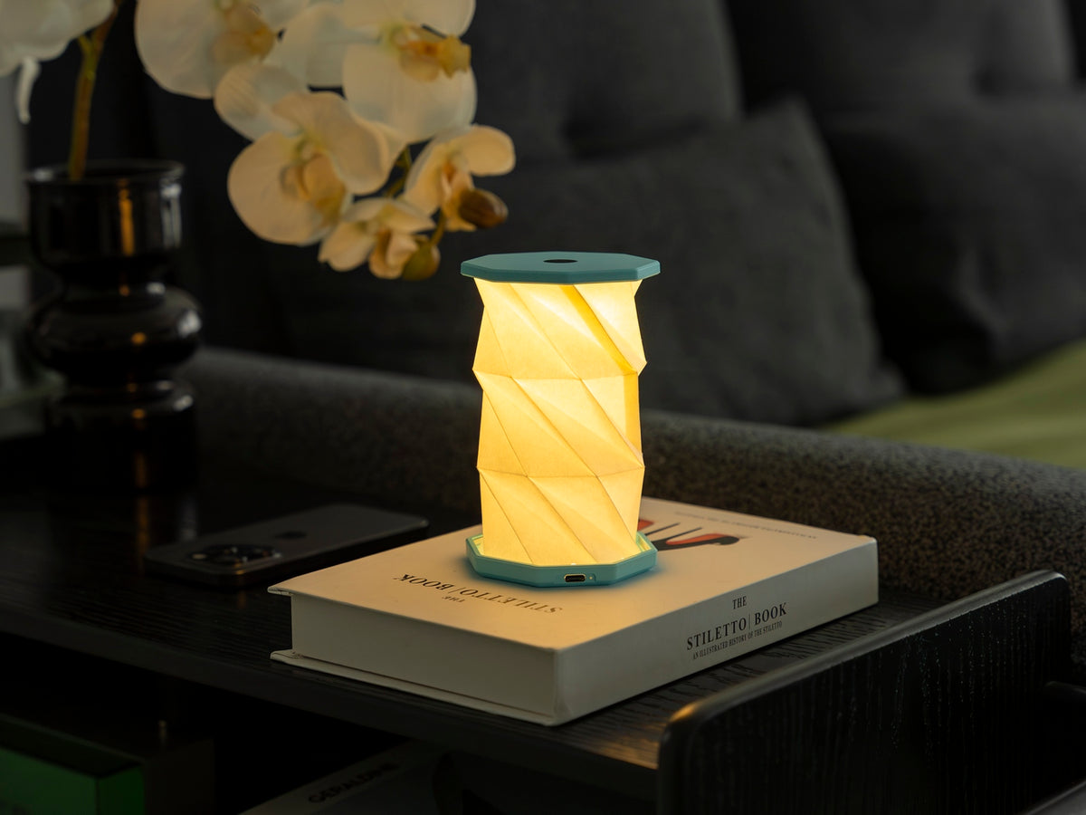 Gingko Design – Luz Twist Hexagon - Azul Laguna - Mini, lámpara de mesa plegable encendida sobre un libro en un salón