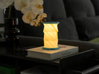 Gingko Design – Luz Twist Hexagon - Azul Laguna - Mini, lámpara de mesa plegable encendida sobre un libro en un salón en penumbra